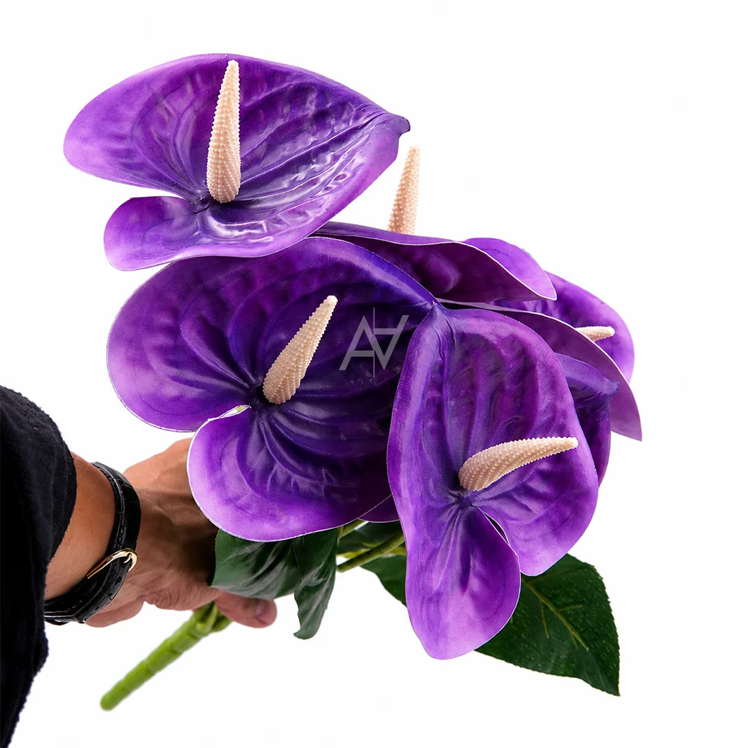 A bouquet of artificial purple anthurium flowers with green leaves and pale spatels, held by a person's hand against a plain white background.
