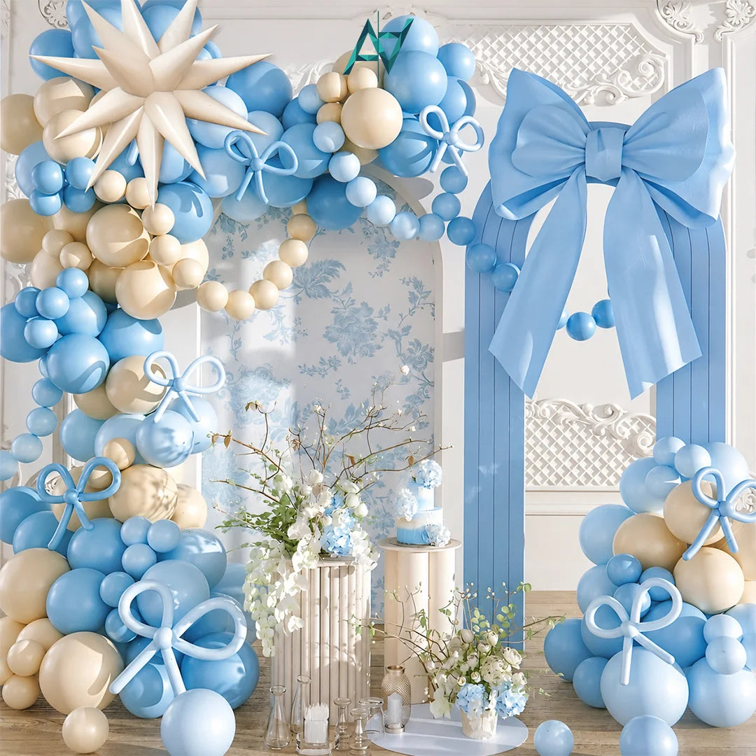A decorative party setup featuring a large balloon arch and matching backdrop in light blue and cream, with a giant blue bow, small gift boxes, floral arrangements, and a blue teddy bear on a pedestal.